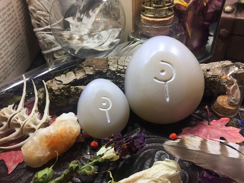 May include: Two white, egg-shaped stones with a carved symbol on each. The stones are sitting on a wooden surface with other decorative items.
