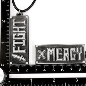 May include: Two metal keychains with a black cord. One keychain has the word "FIGHT" in pixelated letters and the other keychain has the word "MERCY" in pixelated letters.