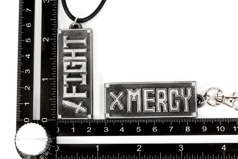 May include: Two metal keychains with a black cord. One keychain has the word "FIGHT" in pixelated letters and the other keychain has the word "MERCY" in pixelated letters.