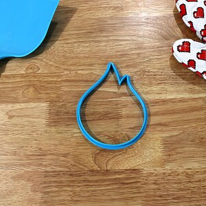 May include: A blue plastic cookie cutter shaped like a flame on a wooden surface.