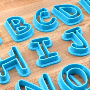 Op de afbeelding: Een set blauwe plastic alfabetkoekjesuitstekers. De uitstekers zijn gerangschikt op een houten oppervlak. De letters zijn A, B, C, D, E, H, I, J, K, N, O en P.