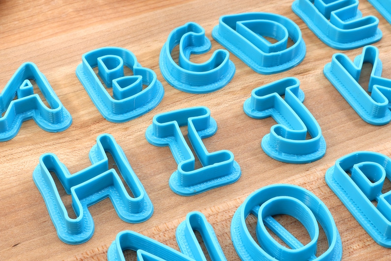 Op de afbeelding: Een set blauwe plastic alfabetkoekjesuitstekers. De uitstekers zijn gerangschikt op een houten oppervlak. De letters zijn A, B, C, D, E, H, I, J, K, N, O en P.