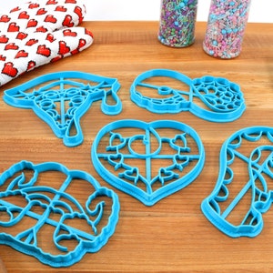 May include: Set of six blue plastic cookie cutters on a wooden surface. The cutters are shaped like a graduation cap, a heart, a donut, a candy cane, and two other shapes.