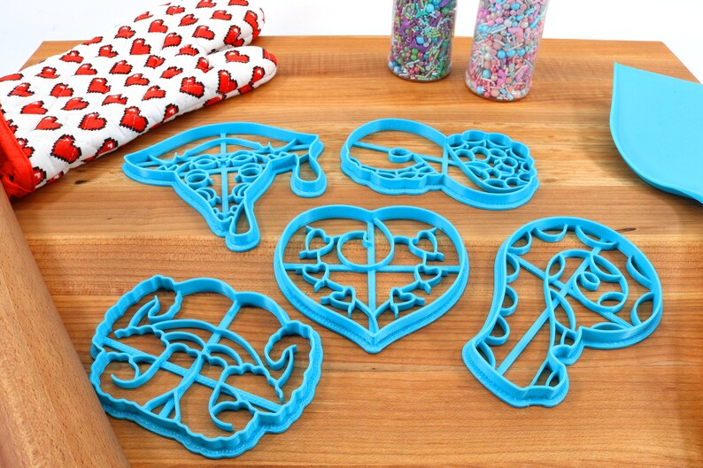 May include: Set of six blue plastic cookie cutters on a wooden surface. The cutters are shaped like a graduation cap, a heart, a donut, a candy cane, and two other shapes.