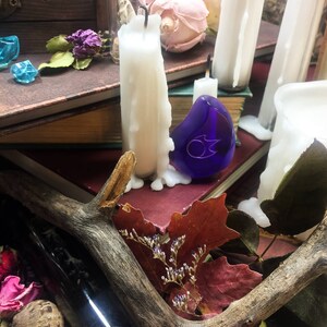 Op de afbeelding: Een blauwe glazen hanger met een zilveren ontwerp ligt op een stapel boeken met witte kaarsen en gedroogde bloemen. Een houten geweistang en een glazen fles bevinden zich op de voorgrond.