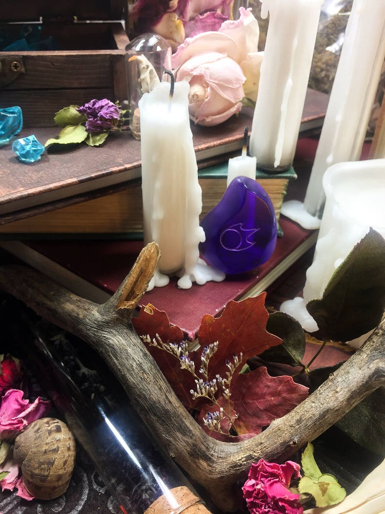 Op de afbeelding: Een blauwe glazen hanger met een zilveren ontwerp ligt op een stapel boeken met witte kaarsen en gedroogde bloemen. Een houten geweistang en een glazen fles bevinden zich op de voorgrond.