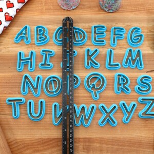 Op de afbeelding: Een set van 26 blauwe plastic alfabetkoekjesuitstekers op een houten oppervlak. De uitstekers zijn alfabetisch gerangschikt. Een liniaal is verticaal in het midden van de afbeelding geplaatst.