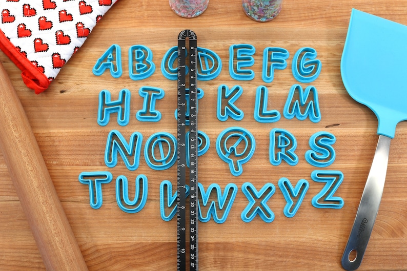 Op de afbeelding: Een set van 26 blauwe plastic alfabetkoekjesuitstekers op een houten oppervlak. De uitstekers zijn alfabetisch gerangschikt. Een liniaal is verticaal in het midden van de afbeelding geplaatst.