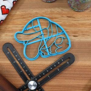 May include: A blue plastic cookie cutter shaped like a cartoon character with a hat. The cutter is on a wooden surface with a ruler and a rolling pin.