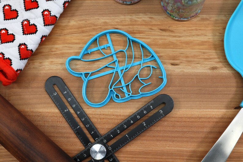May include: A blue plastic cookie cutter shaped like a cartoon character with a hat. The cutter is on a wooden surface with a ruler and a rolling pin.