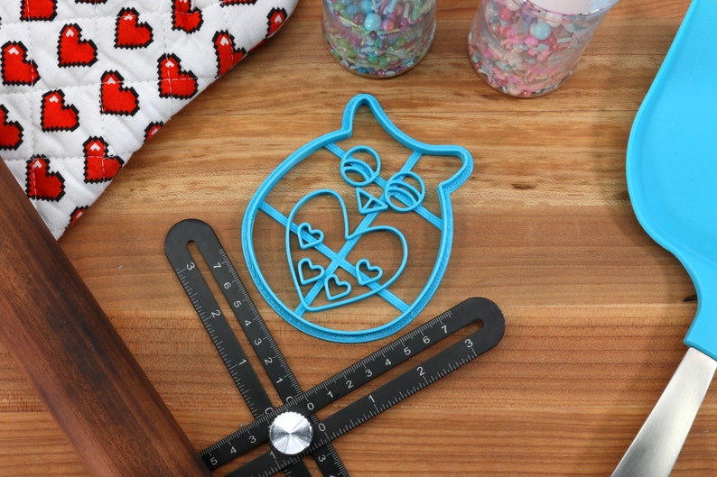 以下が含まれることがあります： ハート型の模様が入ったフクロウの形をした青いプラスチック製のクッキーカッター。カッターは定規と麺棒のある木製の表面の上に置かれています。