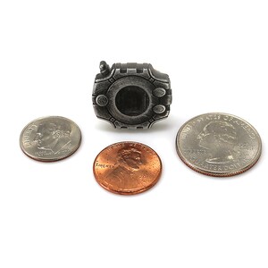 May include: A silver-colored metal replica of a digital pet device, similar in size to a US quarter. The device has a black screen and a raised design around the edges.