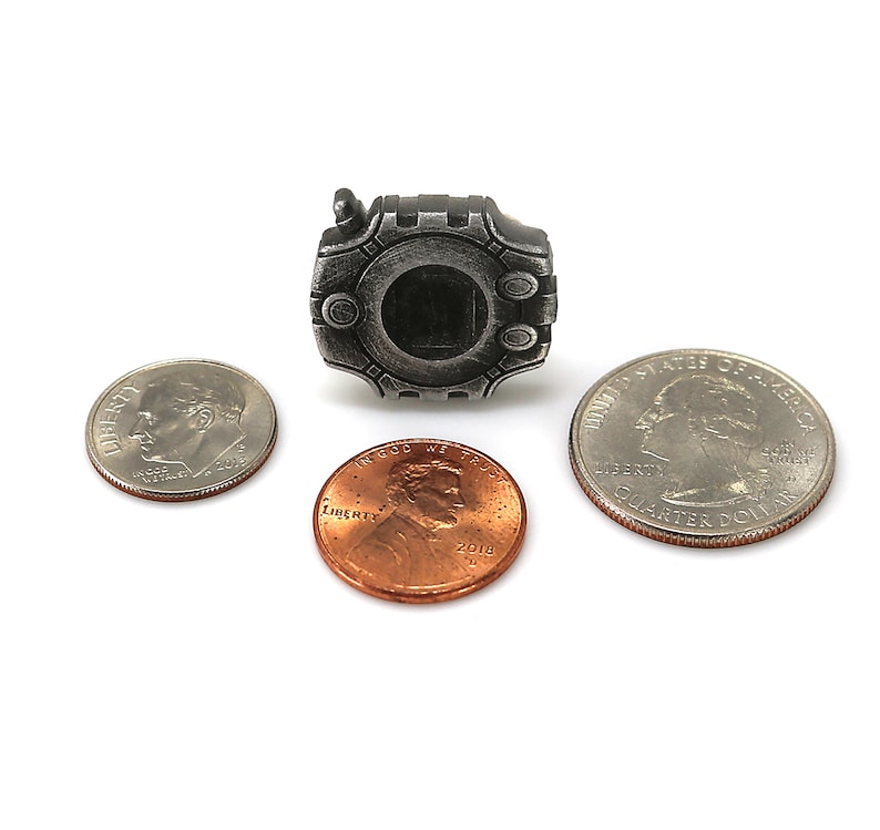 May include: A silver-colored metal replica of a digital pet device, similar in size to a US quarter. The device has a black screen and a raised design around the edges.