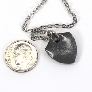 May include: A silver chain necklace with a small, gray, shield-shaped pendant. The pendant has a white marbled design and a silver ring at the top.