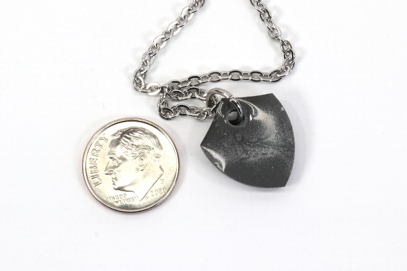 May include: A silver chain necklace with a small, gray, shield-shaped pendant. The pendant has a white marbled design and a silver ring at the top.