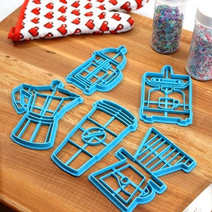 May include: A set of blue plastic cookie cutters shaped like different coffee makers, including a French press, a drip coffee maker, an espresso machine, and a coffee cup. The cutters are on a wooden cutting board with a rolling pin and a spatula.