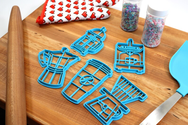 May include: A set of blue plastic cookie cutters shaped like different coffee makers, including a French press, a drip coffee maker, an espresso machine, and a coffee cup. The cutters are on a wooden cutting board with a rolling pin and a spatula.