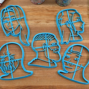 May include: Set of six blue plastic cookie cutters shaped like different meme faces. The cutters are on a wooden surface.