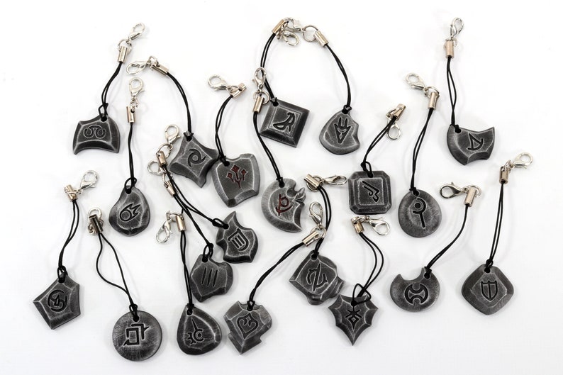 Może przedstawiać: Zestaw 18 srebrnych charm&oacute;w z r&oacute;żnymi symbolami i wzorami. Każdy charm jest przymocowany do czarnego sznurka ze srebrnym zapięciem.