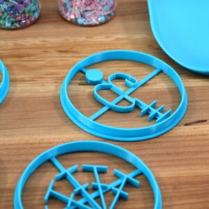 May include: Four blue plastic cookie cutters on a wooden surface. The cutters are shaped like a snowman, a Christmas tree, a snowflake, and a gingerbread man.