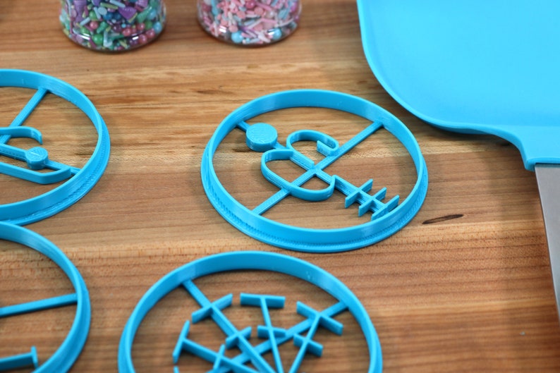 May include: Four blue plastic cookie cutters on a wooden surface. The cutters are shaped like a snowman, a Christmas tree, a snowflake, and a gingerbread man.
