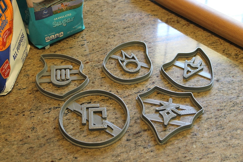 Op de afbeelding: Vijf grijze plastic koekjesvormen met verschillende ontwerpen. De ontwerpen omvatten een ​​muzieknoten, een vlam, een driehoek met een ster erin, een vierkant met een lijn erdoorheen en een Japans karakter.