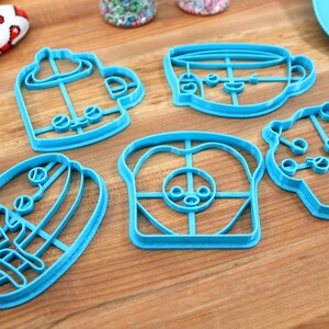 May include: A set of six blue plastic cookie cutters shaped like a coffee cup, a cupcake, a slice of bread, a sandwich, a mug, and a teacup. The cutters are on a wooden surface.