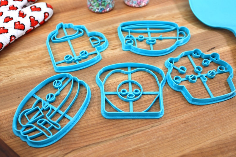 May include: A set of six blue plastic cookie cutters shaped like a coffee cup, a cupcake, a slice of bread, a sandwich, a mug, and a teacup. The cutters are on a wooden surface.