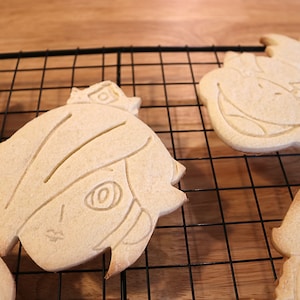 May include: Four sugar cookies on a wire rack. The cookies are shaped like cartoon characters with detailed facial features.