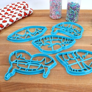 May include: Five blue plastic cookie cutters shaped like a mountain range, a pair of lips, a heart, a pair of eyes, and a mountain range. The cutters are on a wooden surface.