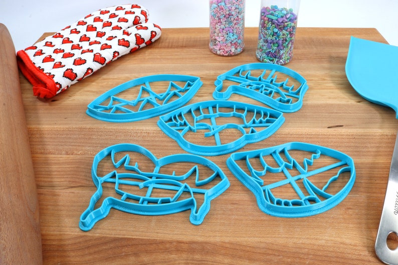 May include: Five blue plastic cookie cutters shaped like a mountain range, a pair of lips, a heart, a pair of eyes, and a mountain range. The cutters are on a wooden surface.