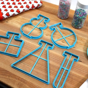May include: A set of blue plastic cookie cutters shaped like a beaker, test tube, graduated cylinder, and a round shape with a cross inside. The cutters are on a wooden surface with a rolling pin, a blue spatula, and two jars of sprinkles.