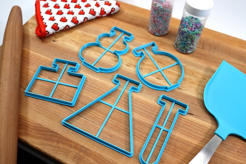 May include: A set of blue plastic cookie cutters shaped like a beaker, test tube, graduated cylinder, and a round shape with a cross inside. The cutters are on a wooden surface with a rolling pin, a blue spatula, and two jars of sprinkles.