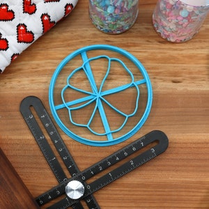 May include: A blue plastic cookie cutter shaped like a flower with five petals. The cutter is on a wooden surface with a black ruler and a rolling pin.