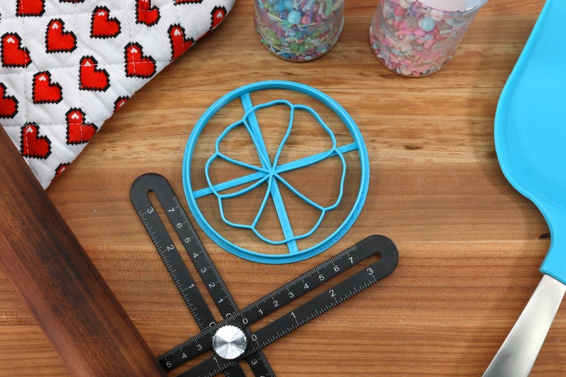 May include: A blue plastic cookie cutter shaped like a flower with five petals. The cutter is on a wooden surface with a black ruler and a rolling pin.