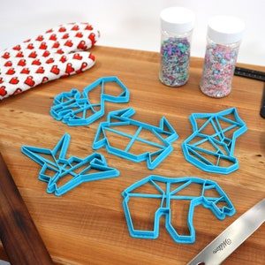 May include: A set of five blue plastic cookie cutters shaped like origami animals, including a crane, a turtle, a bear, a fox, and a deer. The cutters are on a wooden surface with a rolling pin, a spatula, and two jars of sprinkles.