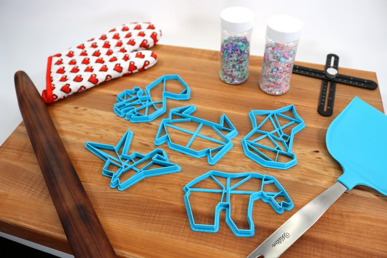 May include: A set of five blue plastic cookie cutters shaped like origami animals, including a crane, a turtle, a bear, a fox, and a deer. The cutters are on a wooden surface with a rolling pin, a spatula, and two jars of sprinkles.