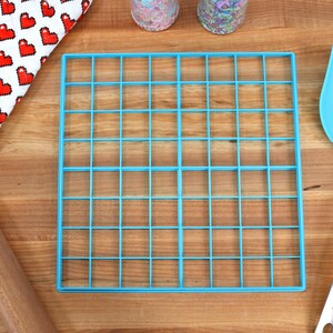 May include: A blue plastic baking grid with 36 squares, a wooden rolling pin, a red and white heart patterned oven mitt, and a blue spatula with a silver handle.