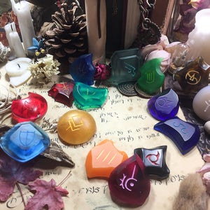 May include: A collection of 14 colorful glass tokens with various symbols and designs. The tokens are arranged on a parchment-like surface with a few decorative items, including a pine cone, candles, and dried flowers.