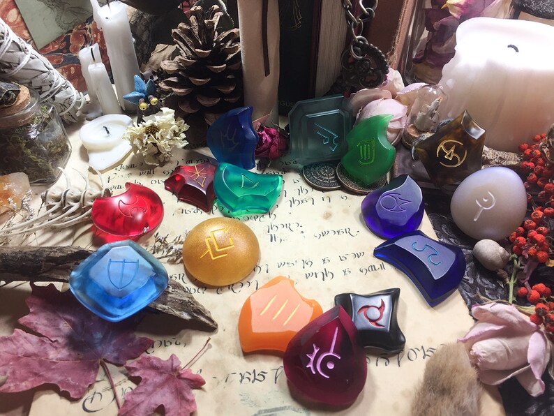May include: A collection of 14 colorful glass tokens with various symbols and designs. The tokens are arranged on a parchment-like surface with a few decorative items, including a pine cone, candles, and dried flowers.