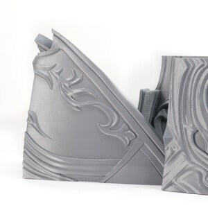 May include: Two gray 3D printed pieces of armor with intricate designs. The pieces are made of a smooth, plastic-like material and have a textured surface.