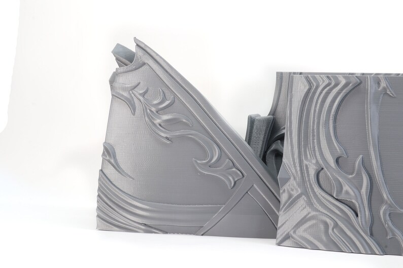 May include: Two gray 3D printed pieces of armor with intricate designs. The pieces are made of a smooth, plastic-like material and have a textured surface.