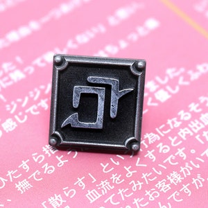 May include: A silver metal pin with a square shape and a black kanji character inside. The pin is on a pink background with Japanese text.