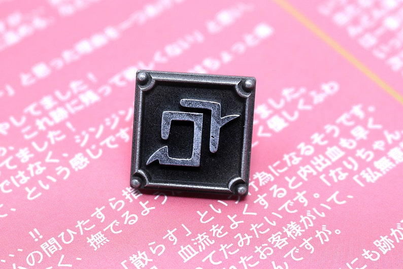 May include: A silver metal pin with a square shape and a black kanji character inside. The pin is on a pink background with Japanese text.