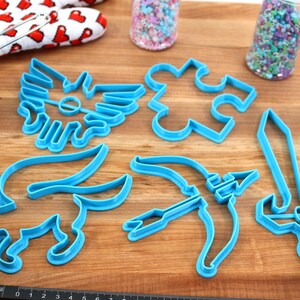 May include: A set of six blue plastic cookie cutters shaped like a sword, a bow and arrow, a puzzle piece, a winged creature, a fox, and a winged symbol. The cutters are on a wooden surface.