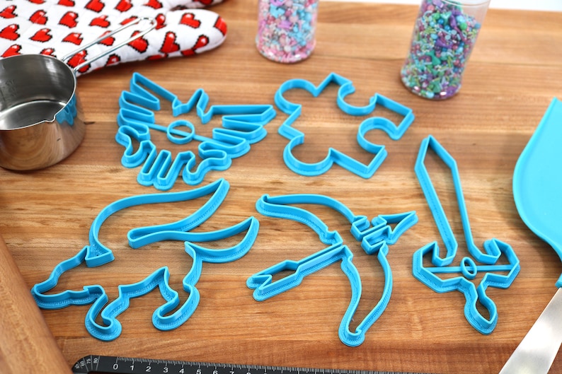 May include: A set of six blue plastic cookie cutters shaped like a sword, a bow and arrow, a puzzle piece, a winged creature, a fox, and a winged symbol. The cutters are on a wooden surface.