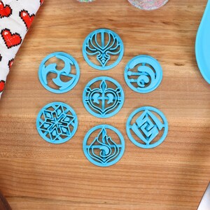 May include: Seven blue plastic cookie cutters with different designs, including a snowflake, a heart, a flame, and a swirl. The cutters are on a wooden surface.