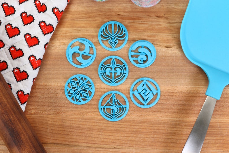 May include: Seven blue plastic cookie cutters with different designs, including a snowflake, a heart, a flame, and a swirl. The cutters are on a wooden surface.