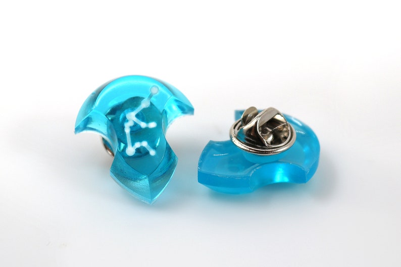 May include: A blue plastic pin with a white constellation design. The pin has a metal clasp on the back.
