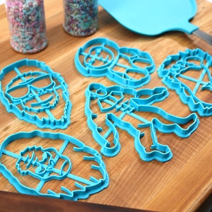May include: A set of six blue plastic cookie cutters shaped like different cartoon characters. The cutters are on a wooden surface with two jars of sprinkles in the background.